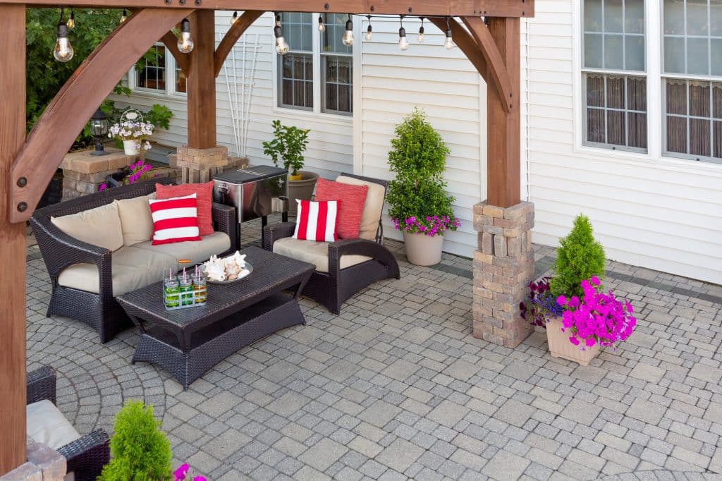 Comfortable chairs with colorful red cushions on an outdoor brick patio covered by a wooden gazebo in front of a timber clad house