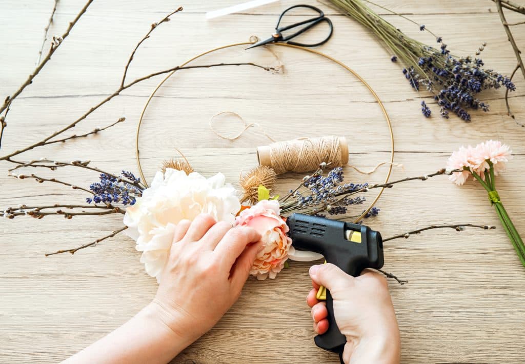 Woman hands making modern spring theme floral arrangement wreath for home decoration. Round metal hoop frame with various real and artificial branches and flowers attached. Catkins, lavender. Hobby.