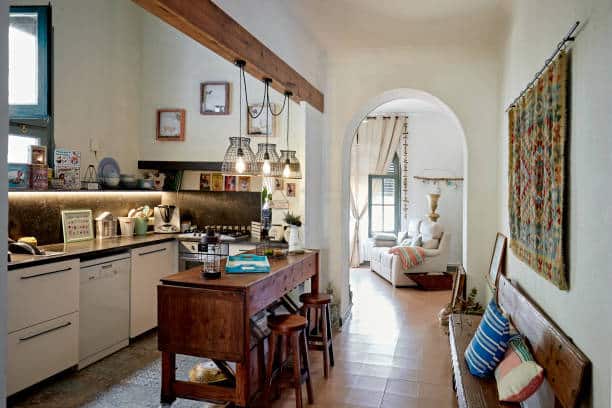 Wide angle view of kitchen with wooden counter-height dining table and stools, hanging light fixtures, bench, and view into living room.