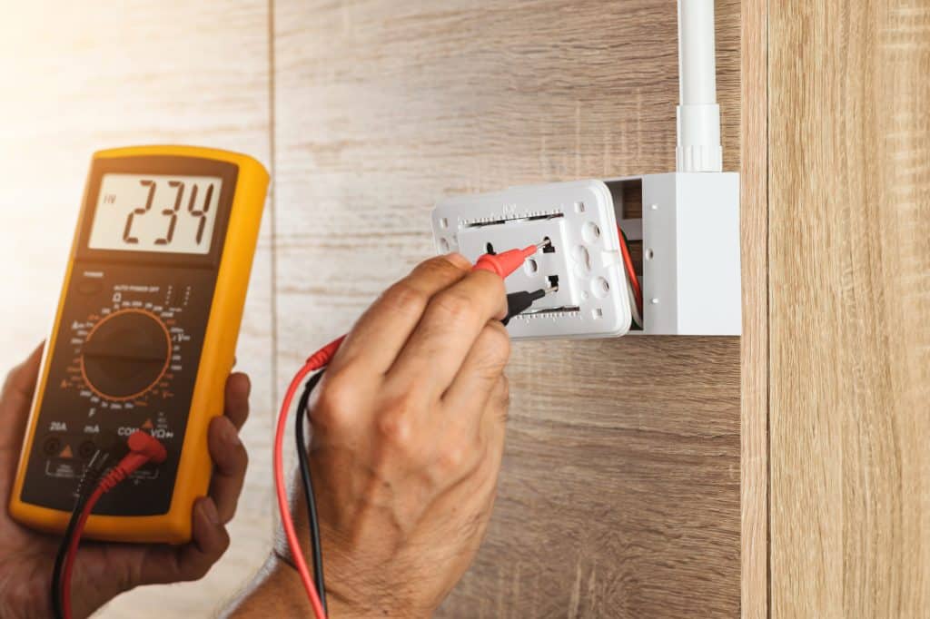 Close up hand of an electrician uses a digital meter to measure the voltage at a wall socket on a wooden wall.